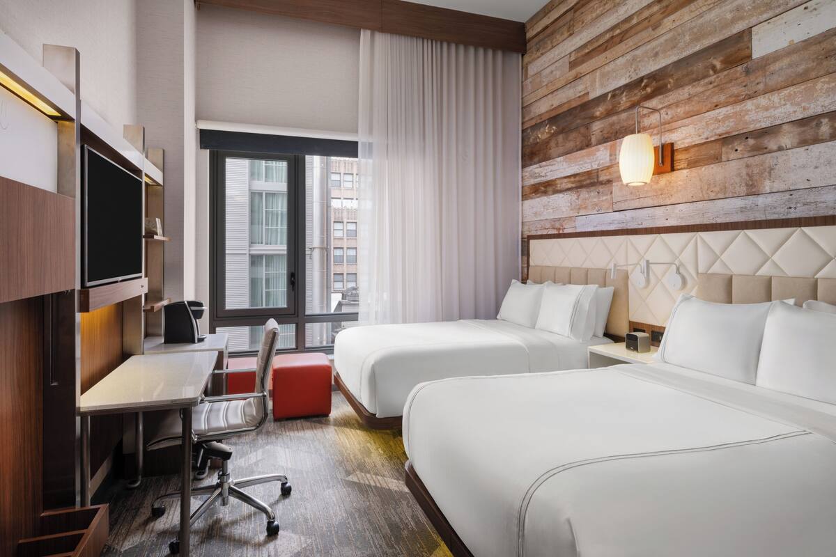 A hotel room with two beds, a desk, a chair, a TV, and a window with curtains.