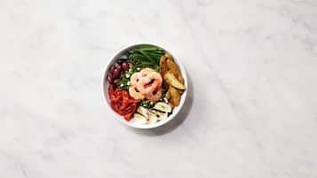 A bowl of food with shrimp, tomatoes, olives, and green beans on a marble surface.