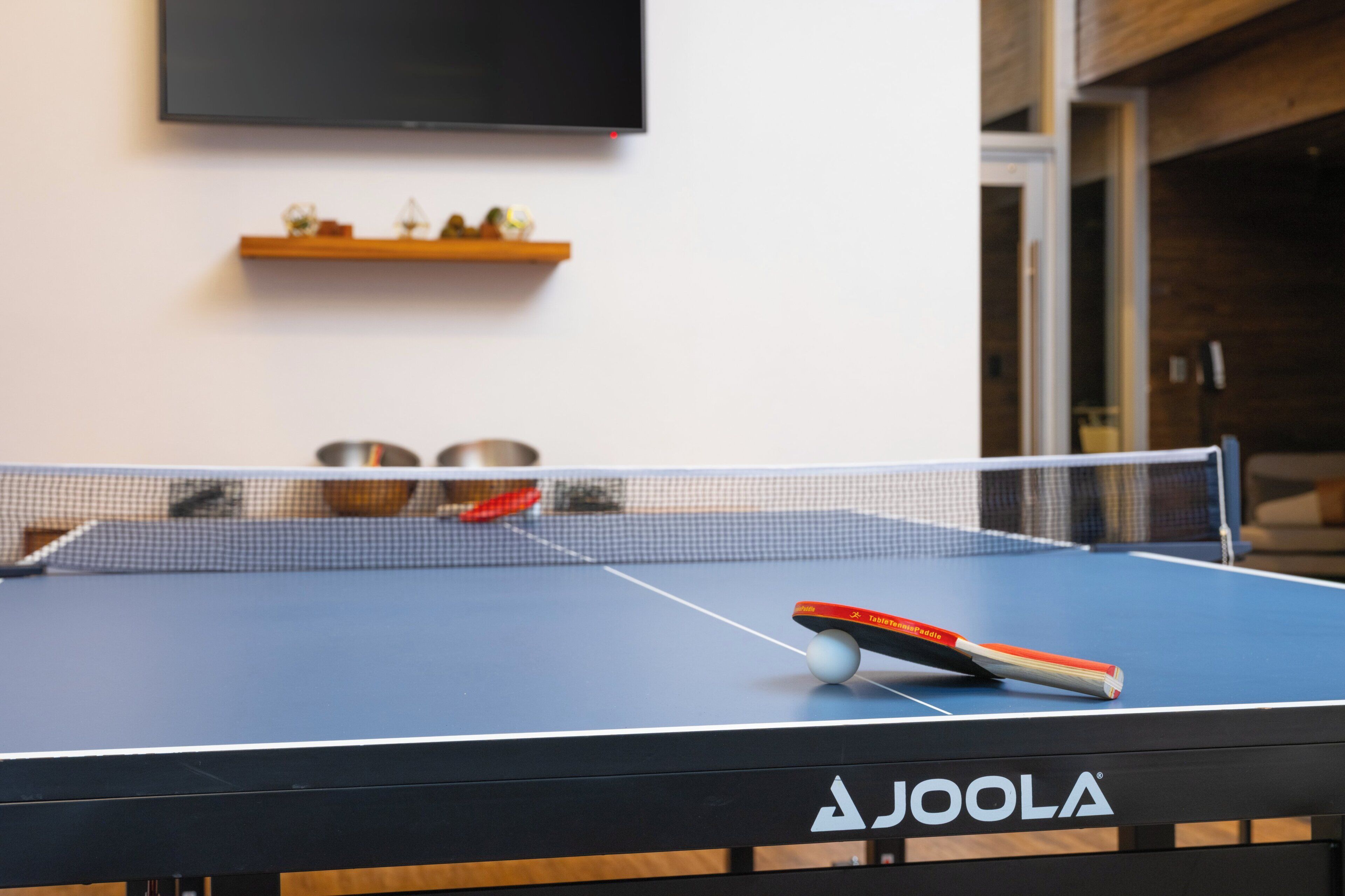 A ping pong table with a net, a TV mounted on the wall, and a shelf with decorative items.