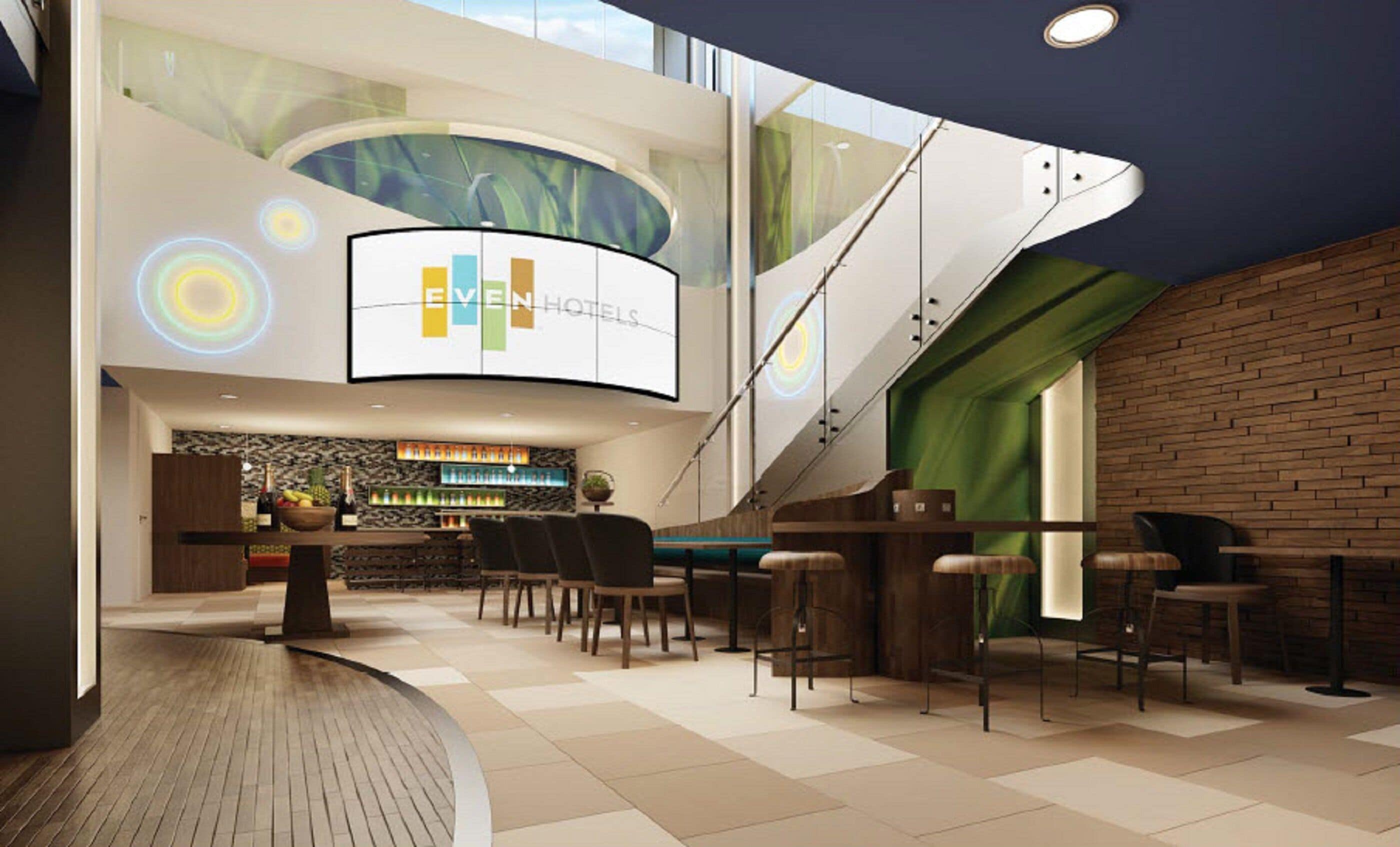 A modern hotel lobby with a curved reception desk, a bar area, and a staircase leading to upper floors.