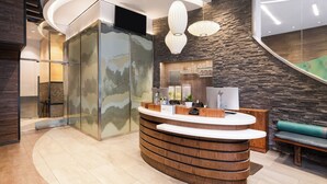 A reception area with a curved wooden counter, a glass wall with a landscape mural, a stone wall, and a bench with a green cushion. - EVEN Hotel New York - Times Square South by IHG (New York)