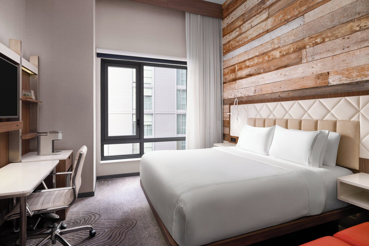 A hotel room with a bed, desk, chair, and a large window.