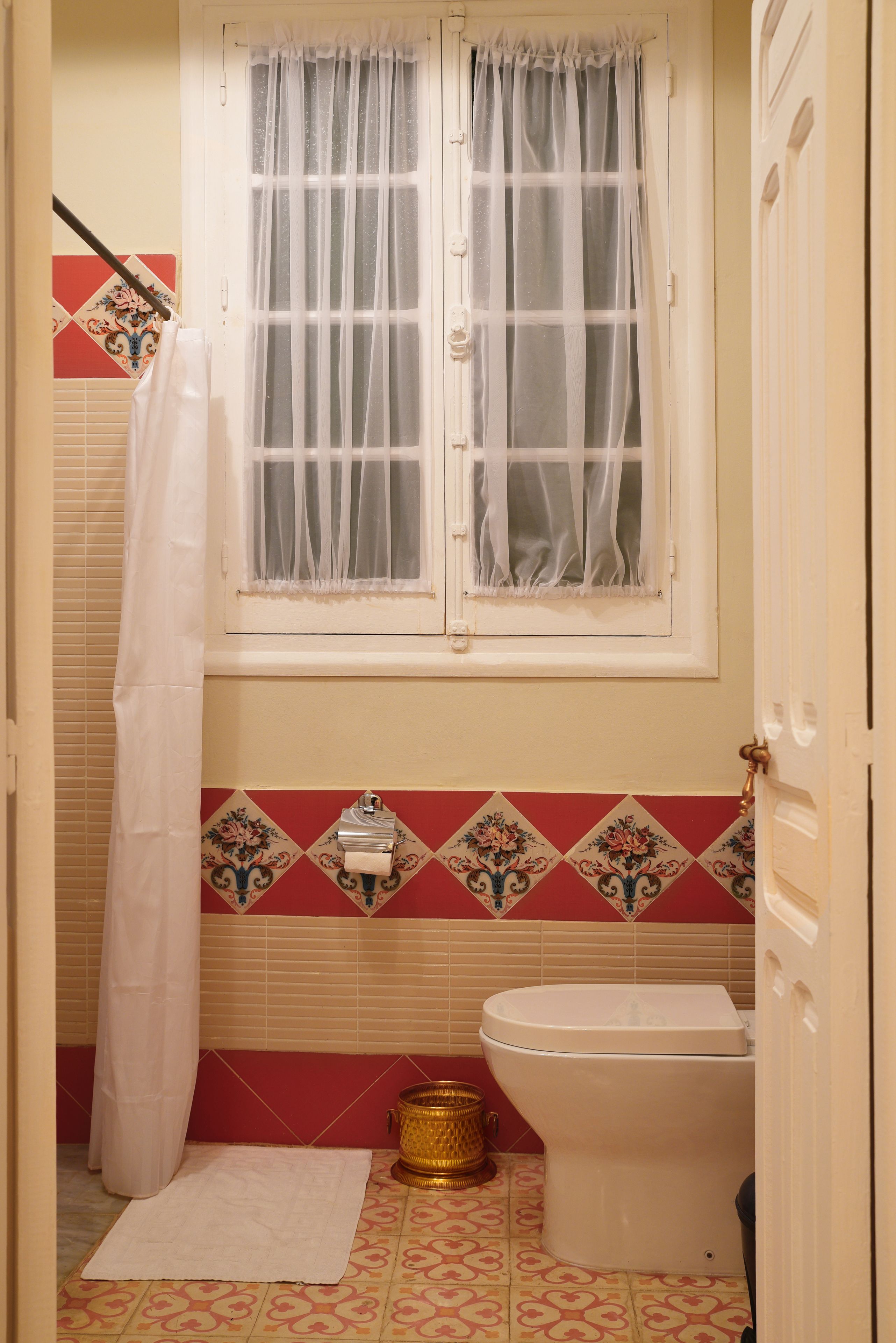 Un baño con inodoro, una ventana con cortinas traslúcidas, una cortina de ducha y azulejos estampados.