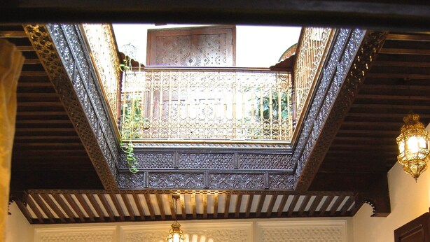 A traditional Moroccan interior with ornate arches, a central courtyard, and a dining area with tables and chairs.