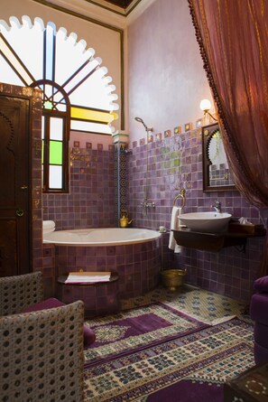 A bathroom with a bathtub, a sink, and a mirror. There is a window with a decorative frame, a rug on the floor, and a curtain on the right side.