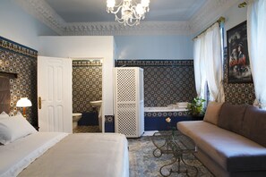 A hotel room with a bed, a sofa, a bathroom with a bathtub, and a chandelier.