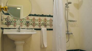 Une salle de bain avec un lavabo blanc, un miroir, une douche équipée d’un pommeau de douche à main et une serviette accrochée au mur.