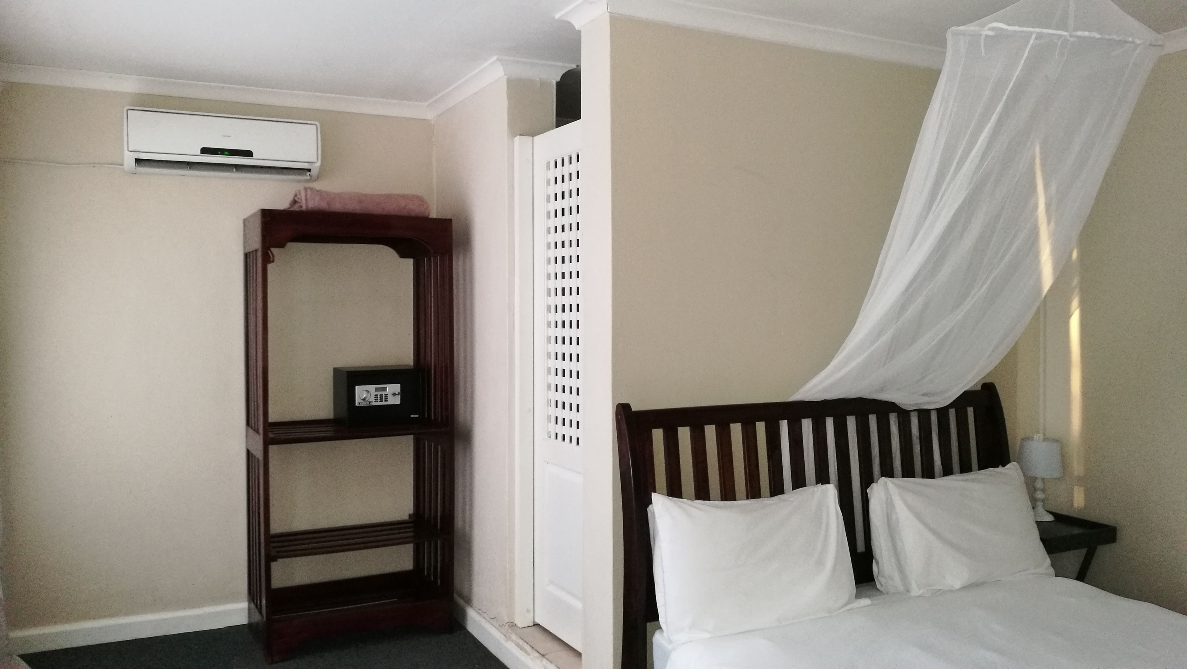 A bedroom with a bed, a nightstand, a bedside table, a wall-mounted air conditioner, a wooden shelf with a safe, and a white canopy over the bed.