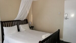 A single bed with a wooden frame and white bedding, a bedside table with a lamp, and a door with a handle. - LePatino Bed & Breakfast (Livingstone)