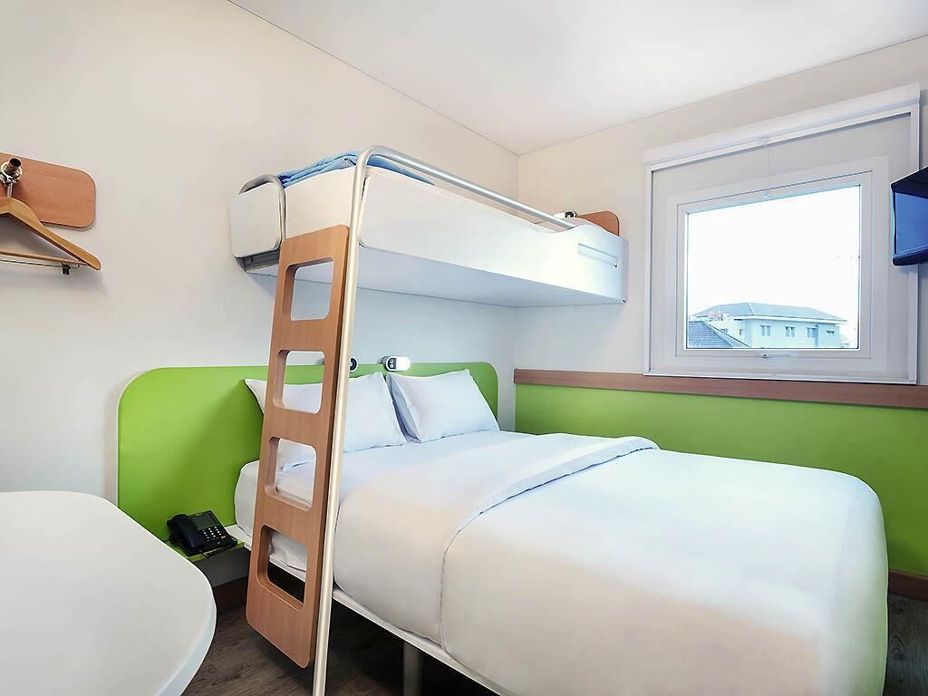 A bunk bed with a ladder, a bedside telephone, and a window with a view of buildings.