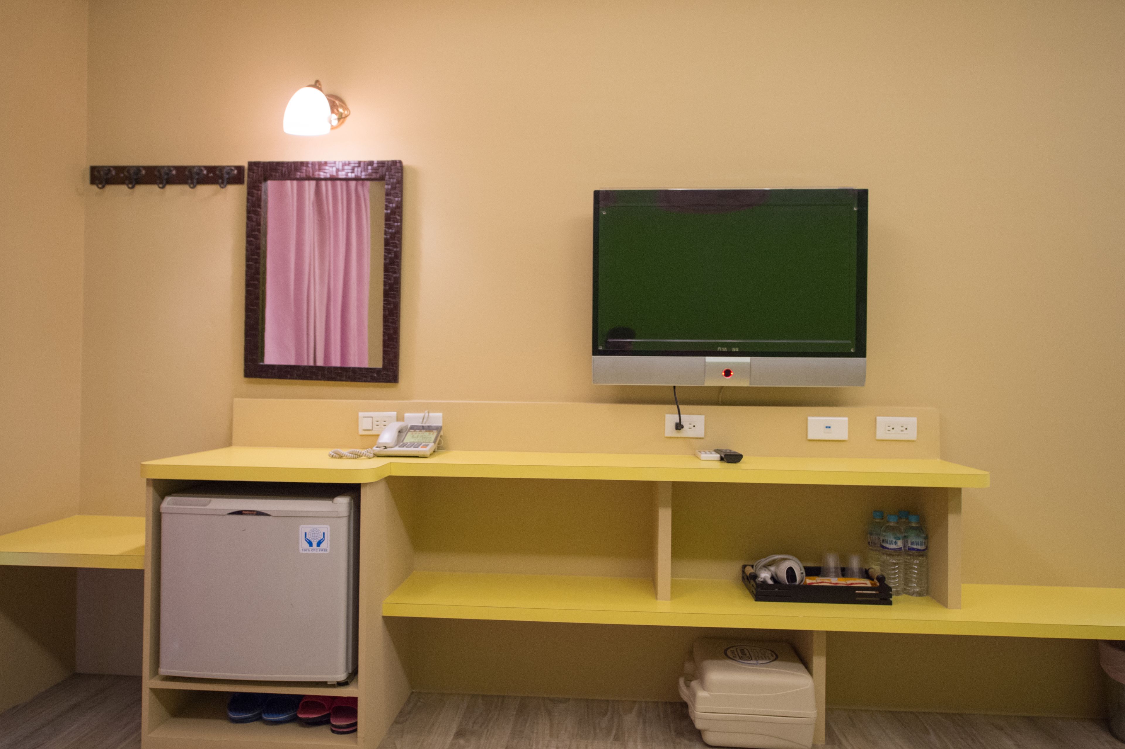 Une petite pièce avec un bureau jaune, un réfrigérateur blanc, une télévision et un miroir surmonté d’un rideau rose.