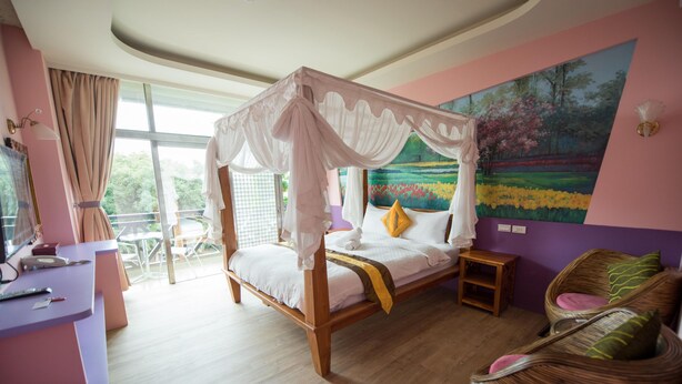 A bedroom with a four-poster bed, a small bedside table, a wicker chair, and a painting on the wall.