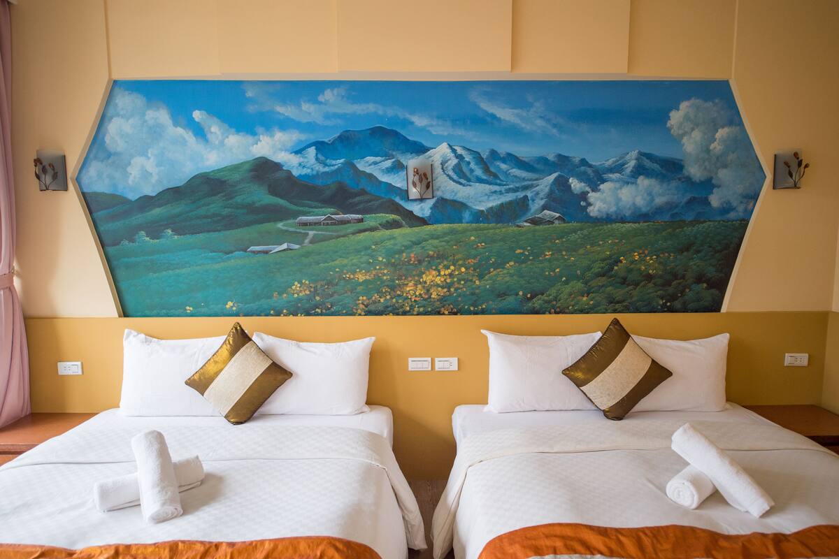 A hotel room with two beds, each with white bedding and a rolled towel. A large painting of a mountainous landscape is on the wall above the beds.