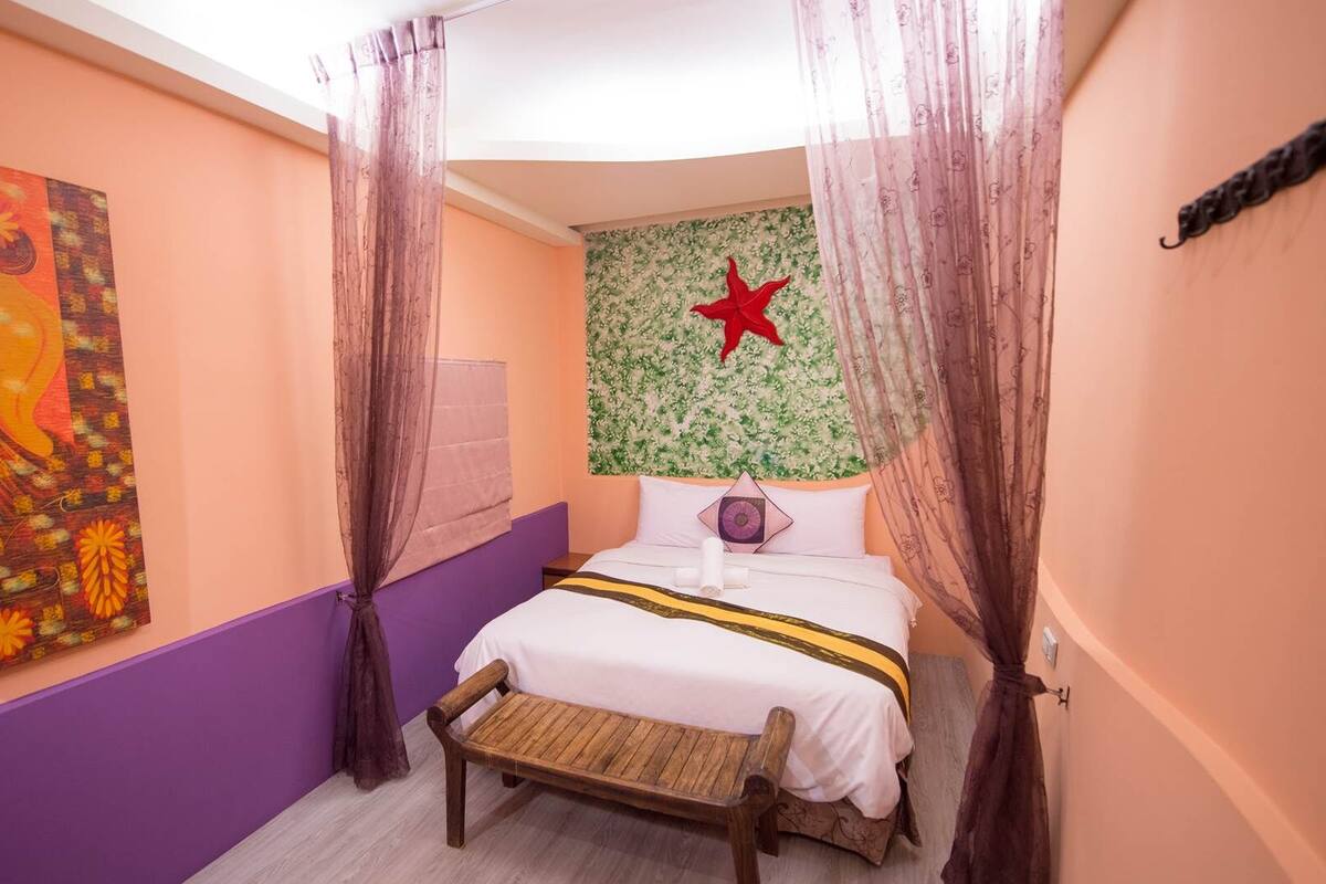 A bedroom with a bed, a bench, a wall with a floral tapestry, and a painting.