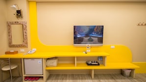 A room with a yellow desk, a television, a mirror, a small refrigerator, and a chair.