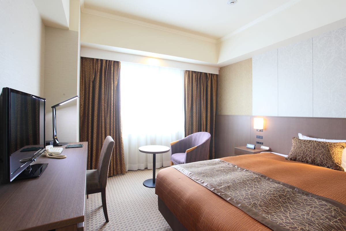 A hotel room with a bed, a desk with a computer, a chair, a small table, and a nightstand with a lamp.