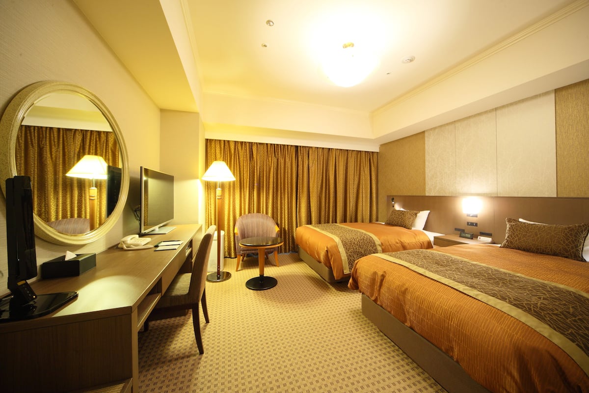 A hotel room with a desk, chair, two beds, a TV, and a large mirror.