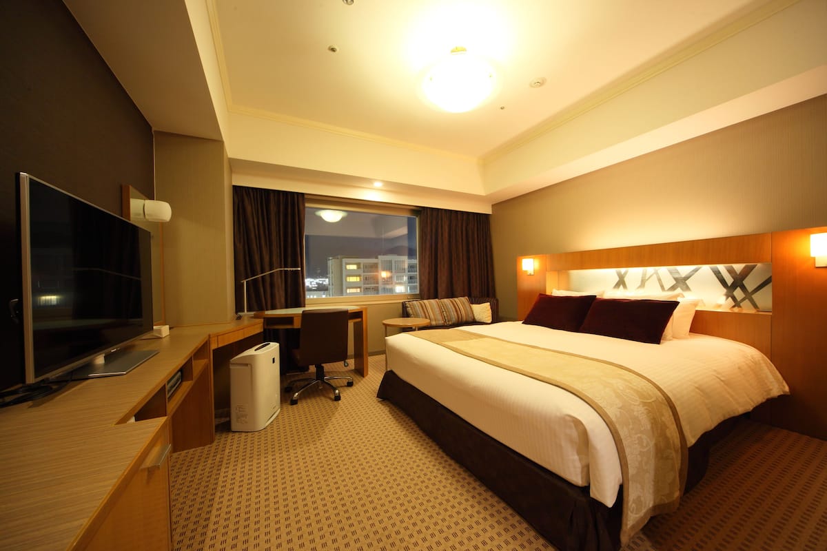A hotel room with a large bed, a desk with a computer, a TV, and a window with curtains.