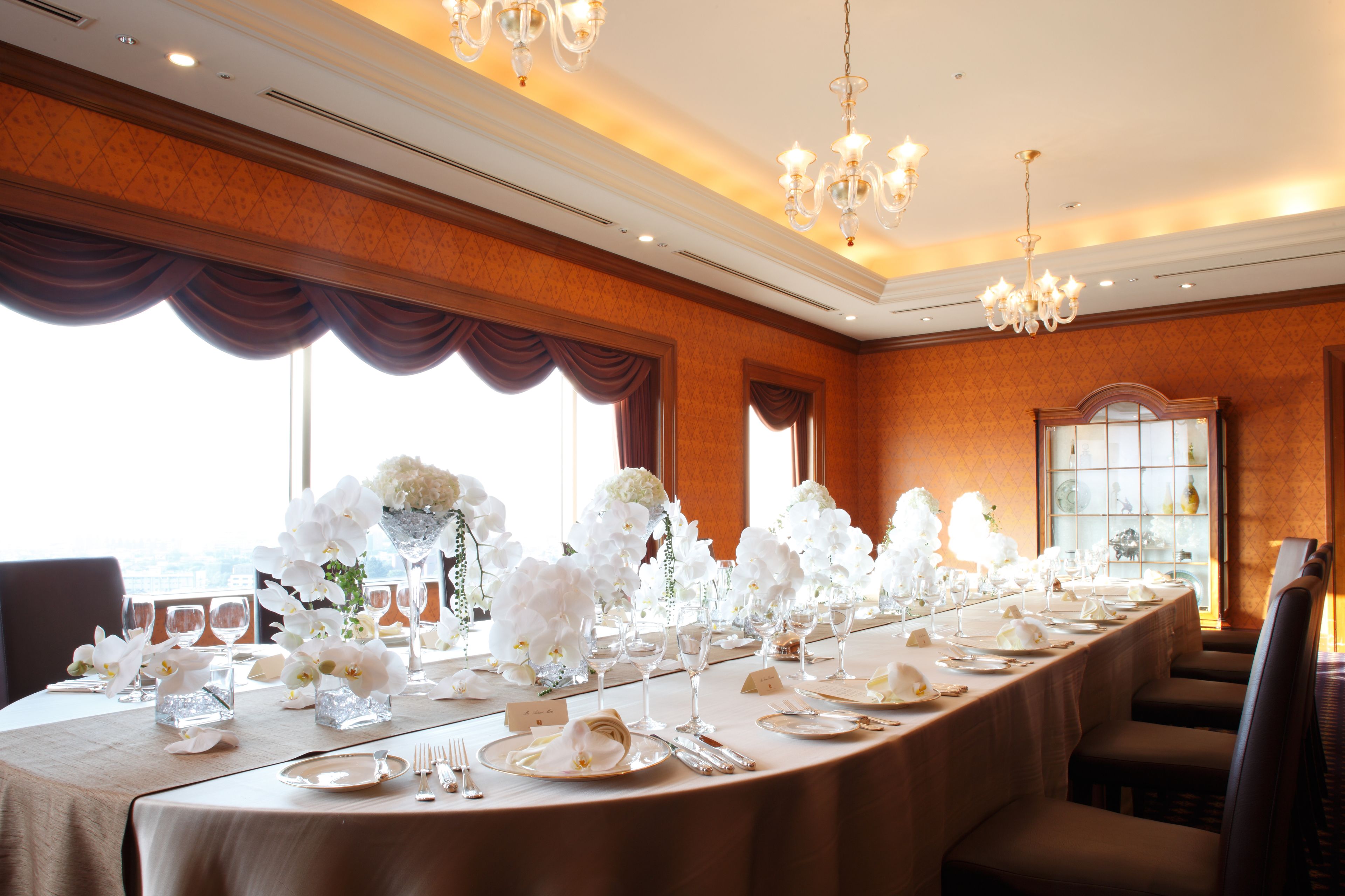 A banquet hall with a long table set for a formal event, featuring white floral centerpieces and elegant table settings.