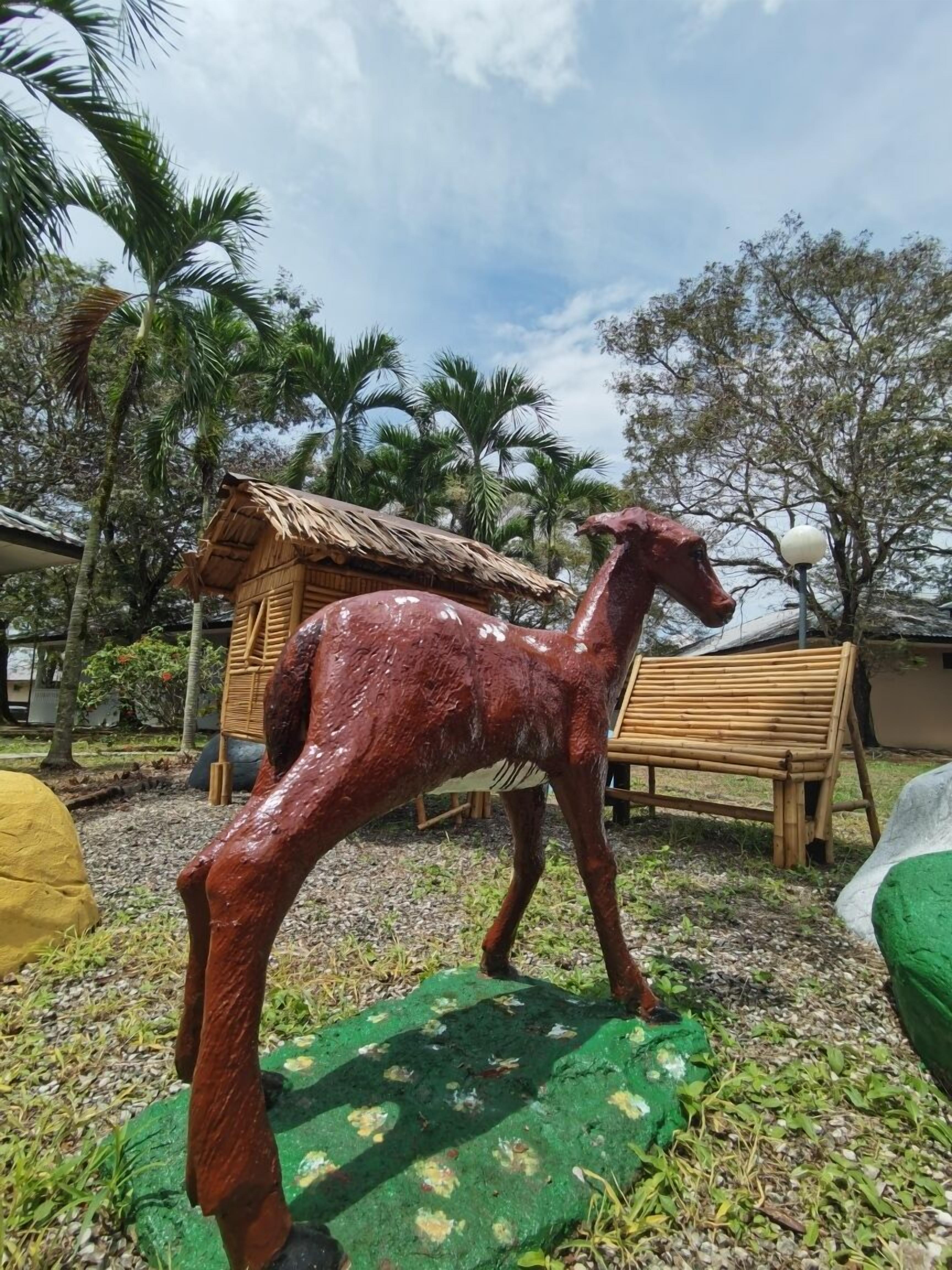 A wooden sculpture of a deer on a green platform, surrounded by palm trees and a thatched-roof structure.
