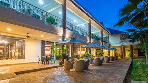 Un patio exterior con mobiliario y sombrillas, un paseo cubierto y un edificio de varios pisos con balcones.