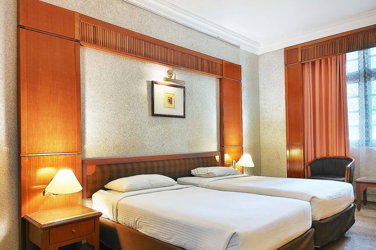 Une chambre d’hôtel avec deux lits, des têtes de lit en bois, un tableau encadré au mur et une fenêtre avec des rideaux.