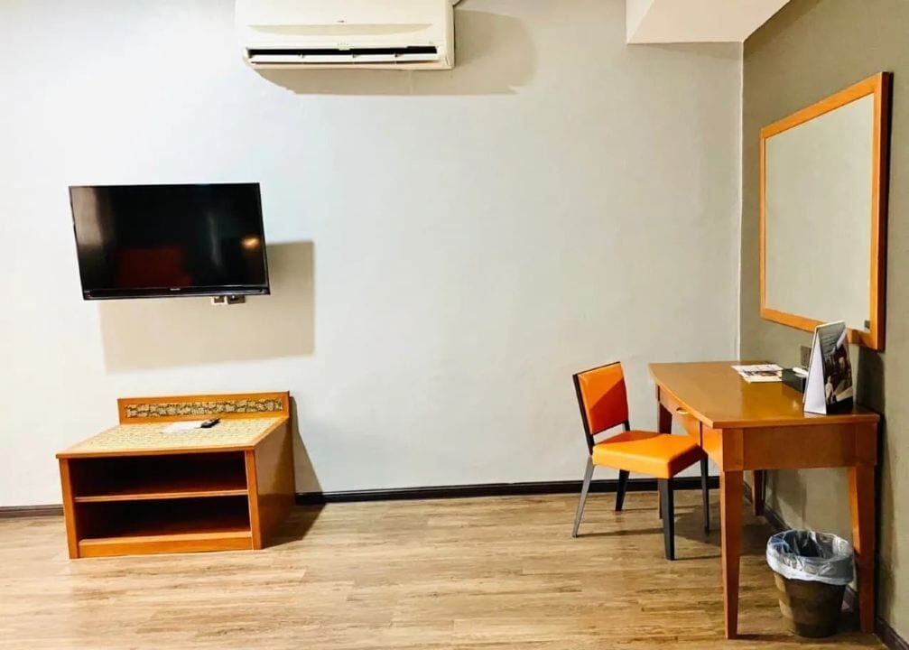 A room with a wooden floor, a TV mounted on the wall, a small wooden table, a chair, and a trash bin.