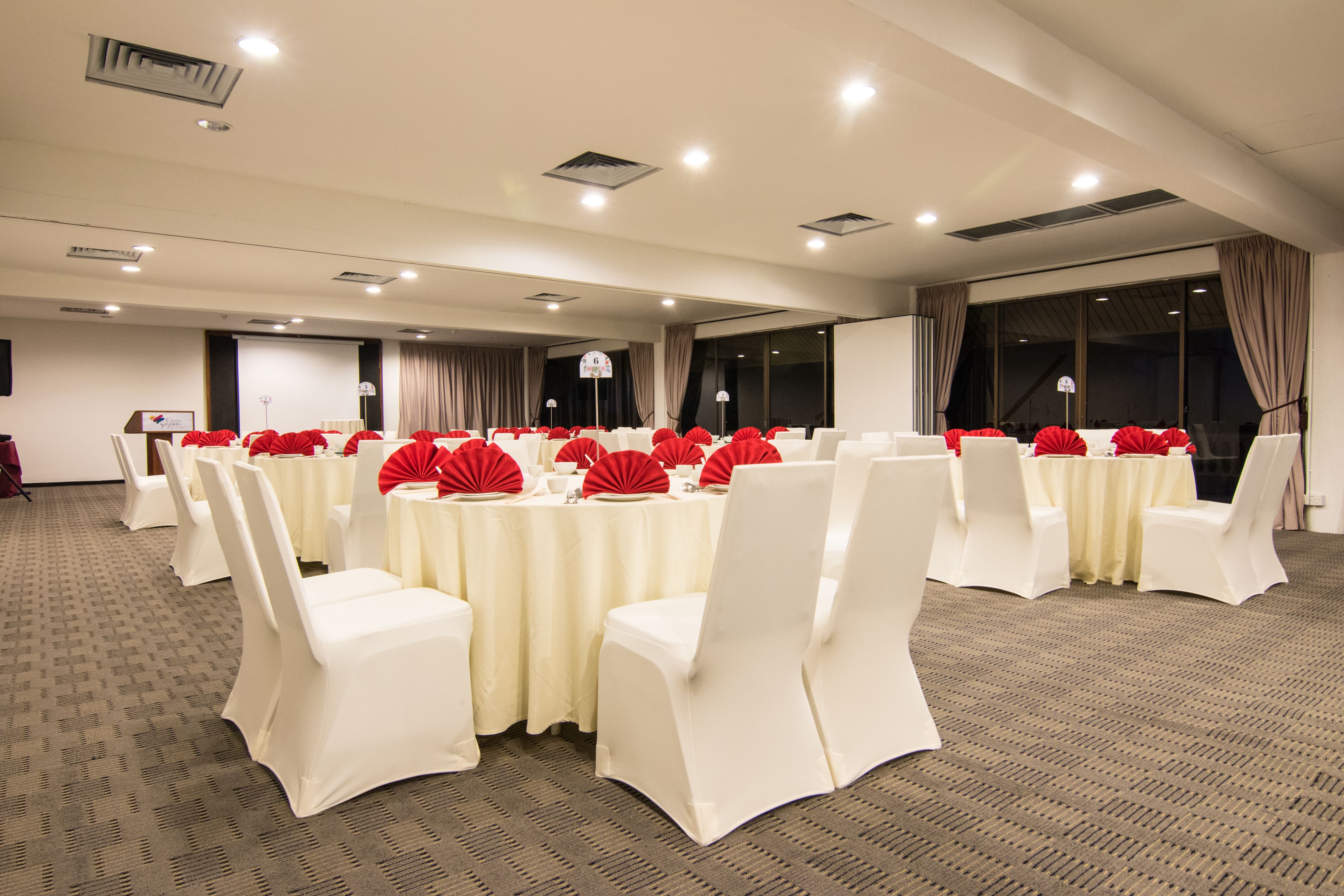 Un salón de conferencias con mesas redondas, sillas blancas y adornos rojos sobre las mesas.