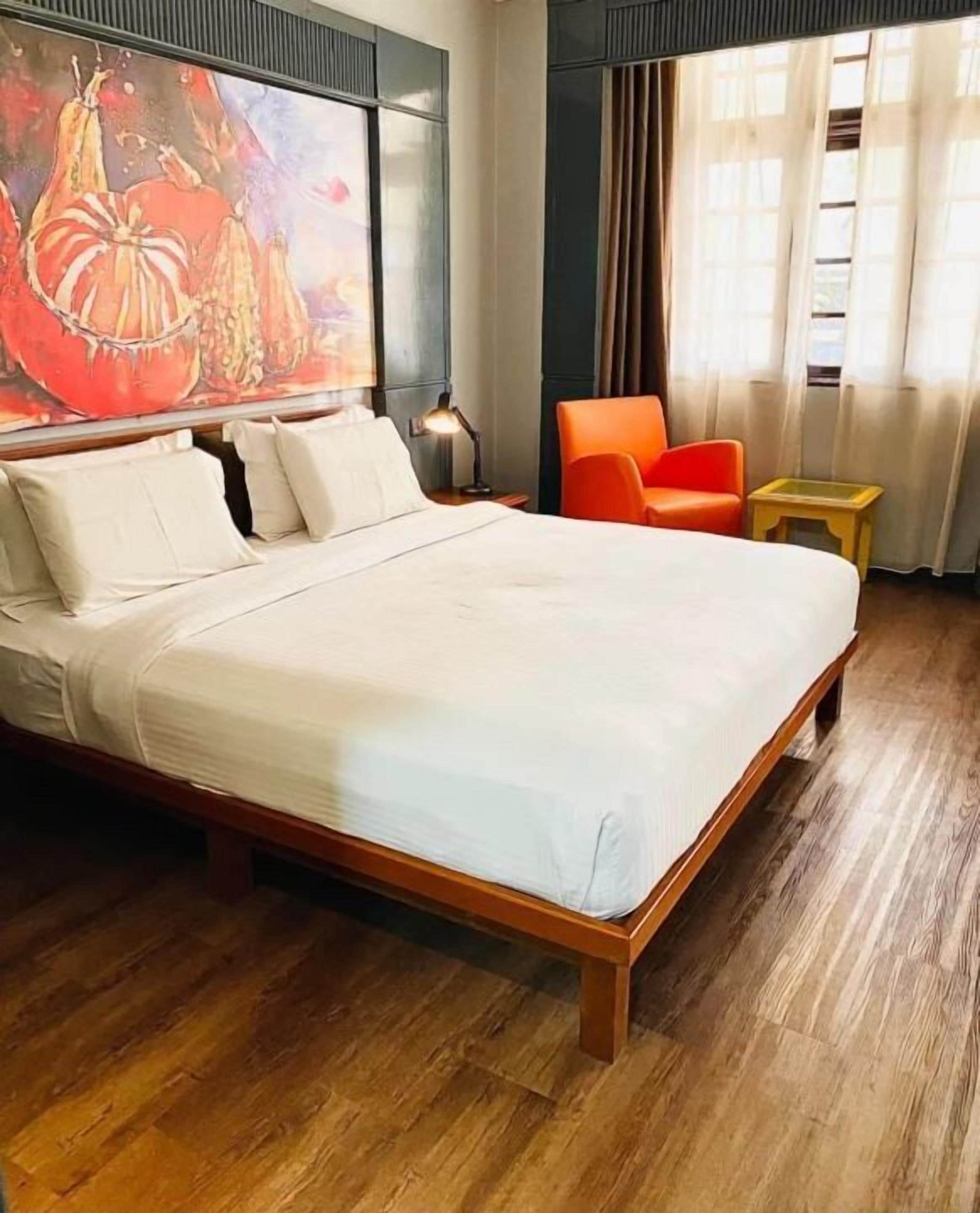 A neatly made bed with white linens, a wooden headboard, and a large abstract painting above it.