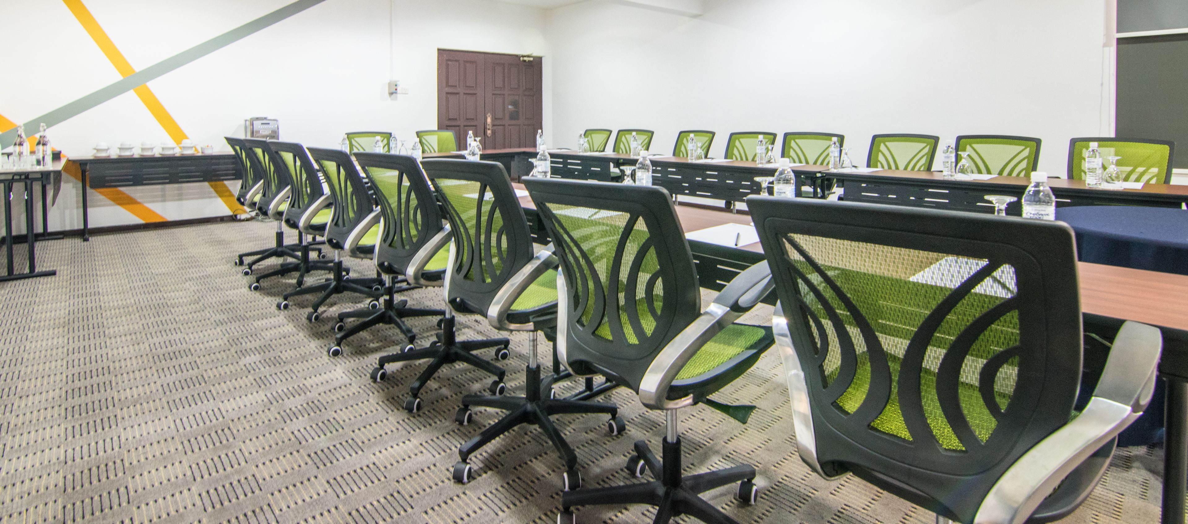 A conference room with rows of green chairs and a long table, equipped with water bottles and a projector.