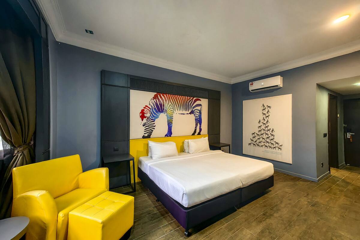 Une chambre d’hôtel moderne avec un grand lit, un fauteuil jaune, une petite table et deux œuvres d’art murales.