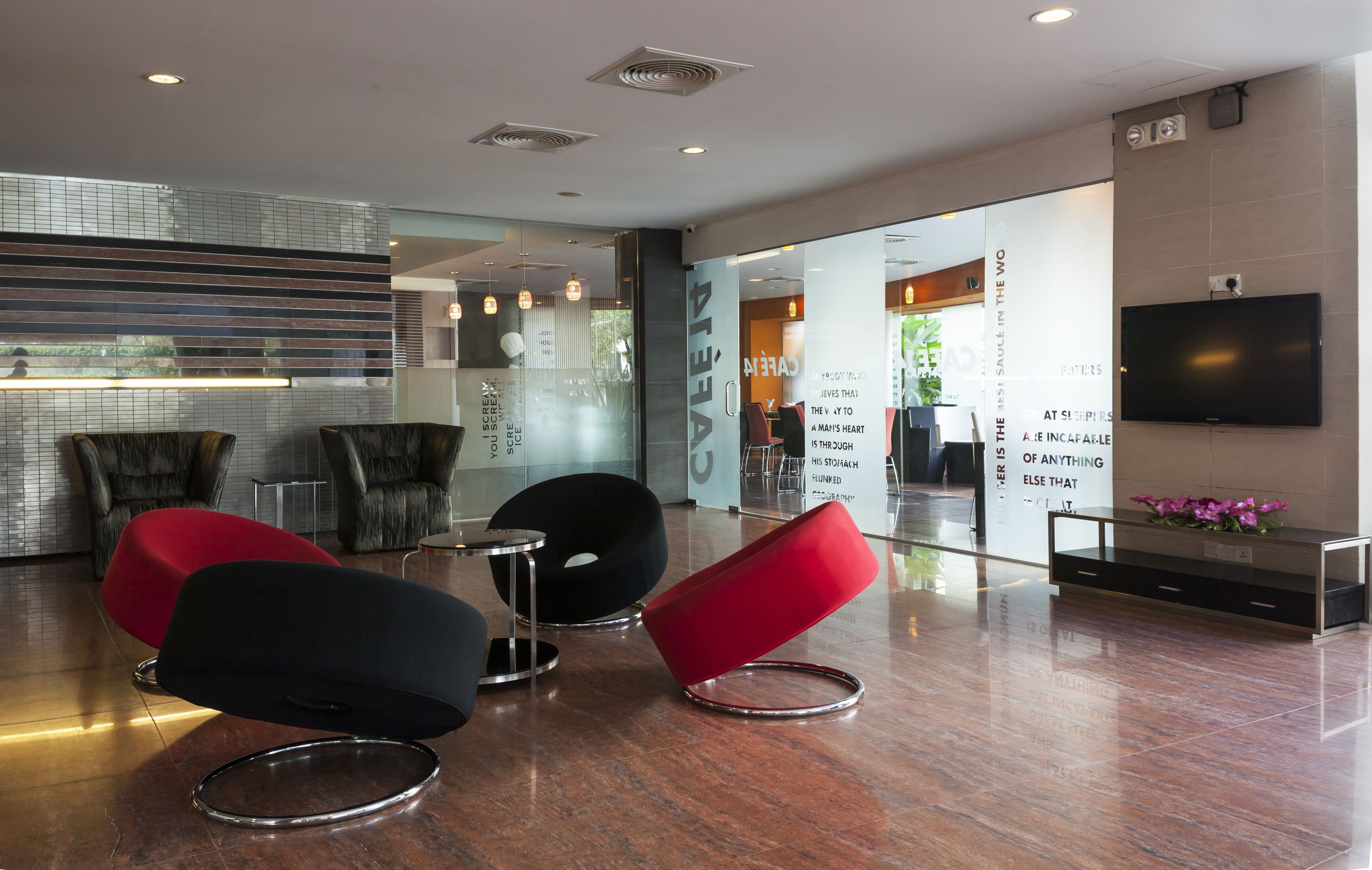 A modern lobby with a glass partition, comfortable seating, a flat-screen TV, and a visible kitchen area.