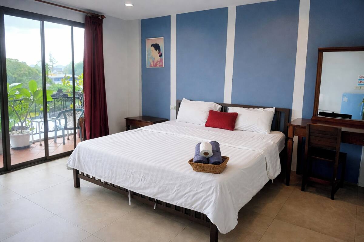 Een hotelkamer met een groot bed, een houten nachtkastje, een bureau en een stoel. Er is een balkon met uitzicht op groen en gebouwen.