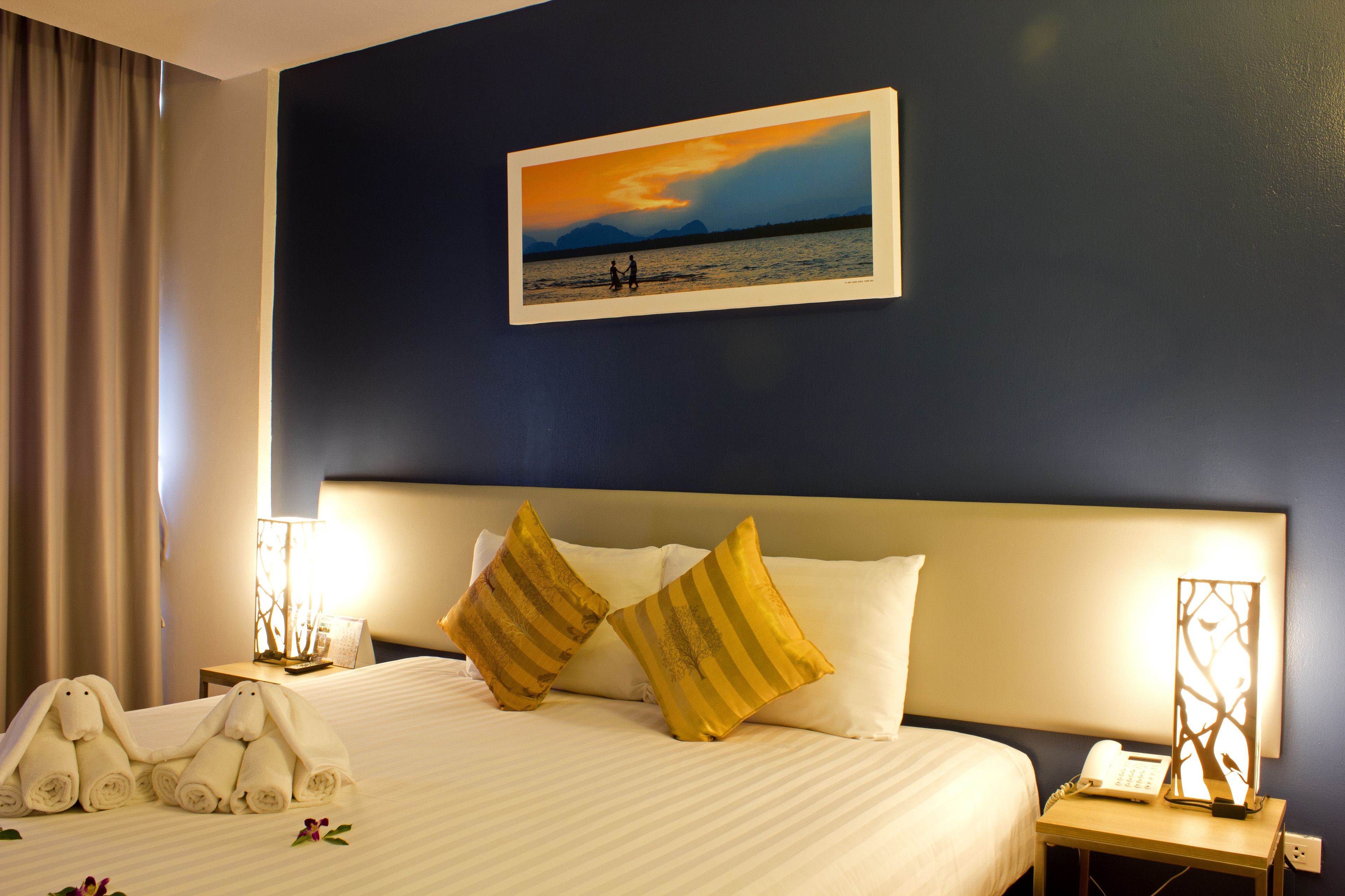 A hotel room with a bed, two pillows, a nightstand with a lamp, a framed picture on the wall, and a window with curtains.