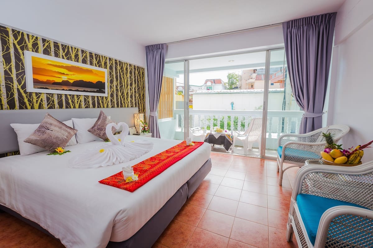 A hotel room with a bed, a balcony with chairs, and a view of buildings.
