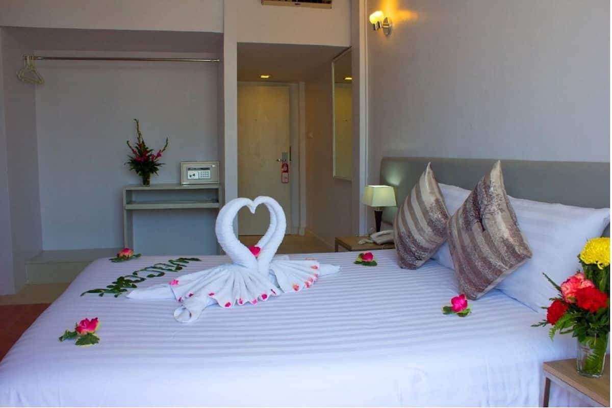 A hotel room with a neatly made bed, a heart-shaped towel decoration, a bedside table with a lamp, and a small vase with flowers.