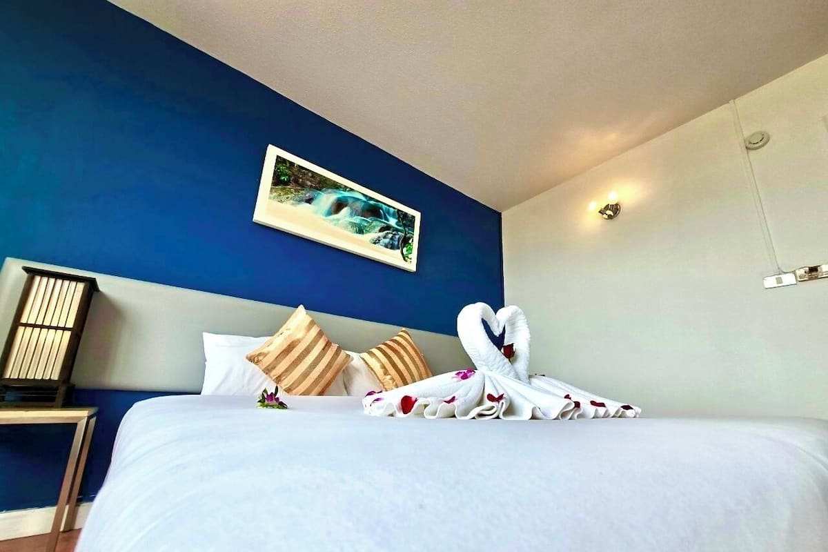 A hotel room with a large bed, blue walls, a painting above the bed, and a wall-mounted light fixture.