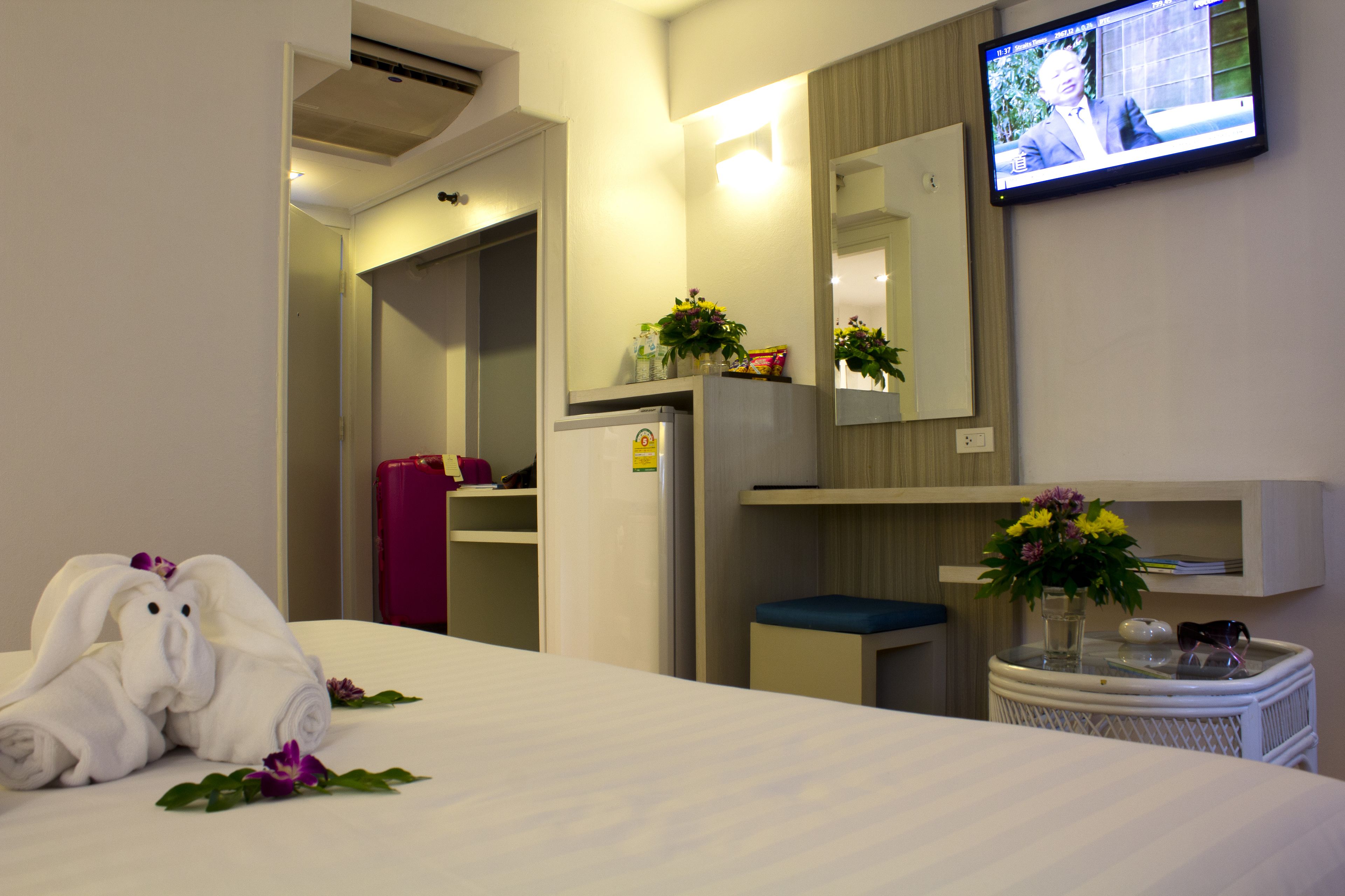 Hotelkamer met een bed, een aan de muur bevestigde televisie, een spiegel, een kleintje tafeltje met een bloevas en een koffer.