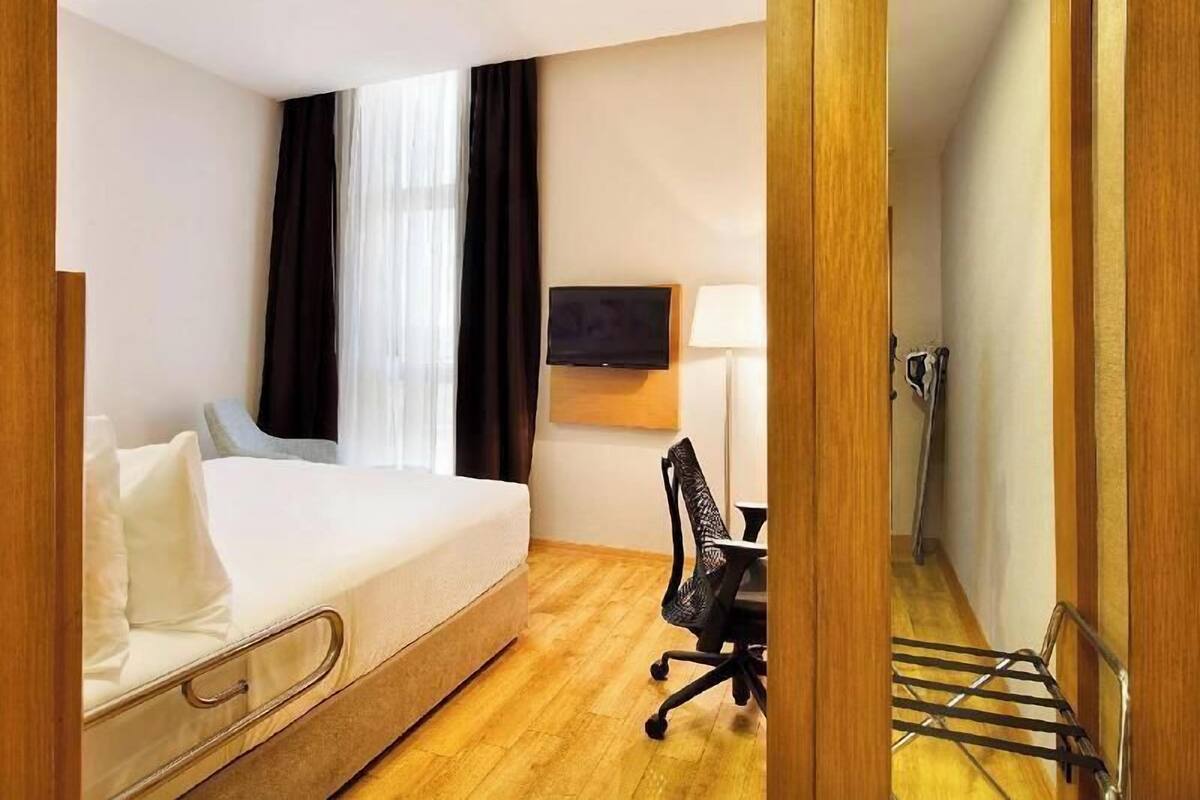 A hotel room with a bed, a desk, a chair, a TV, and a window with curtains.