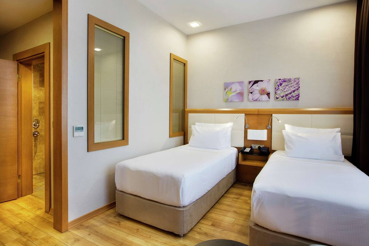 A hotel room with two beds, a wooden floor, a window with blinds, and a wall-mounted artwork.