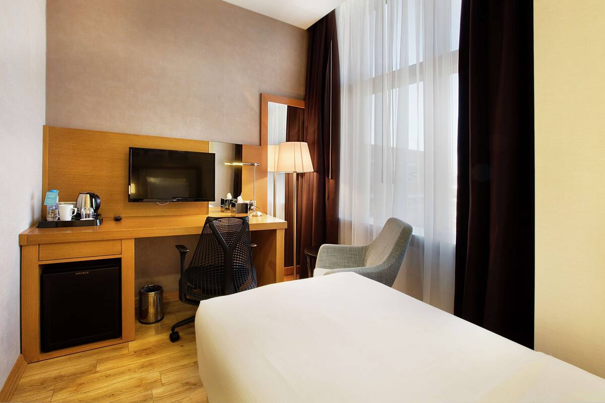 A hotel room with a bed, a desk with a TV, a chair, a lamp, and a window with curtains.