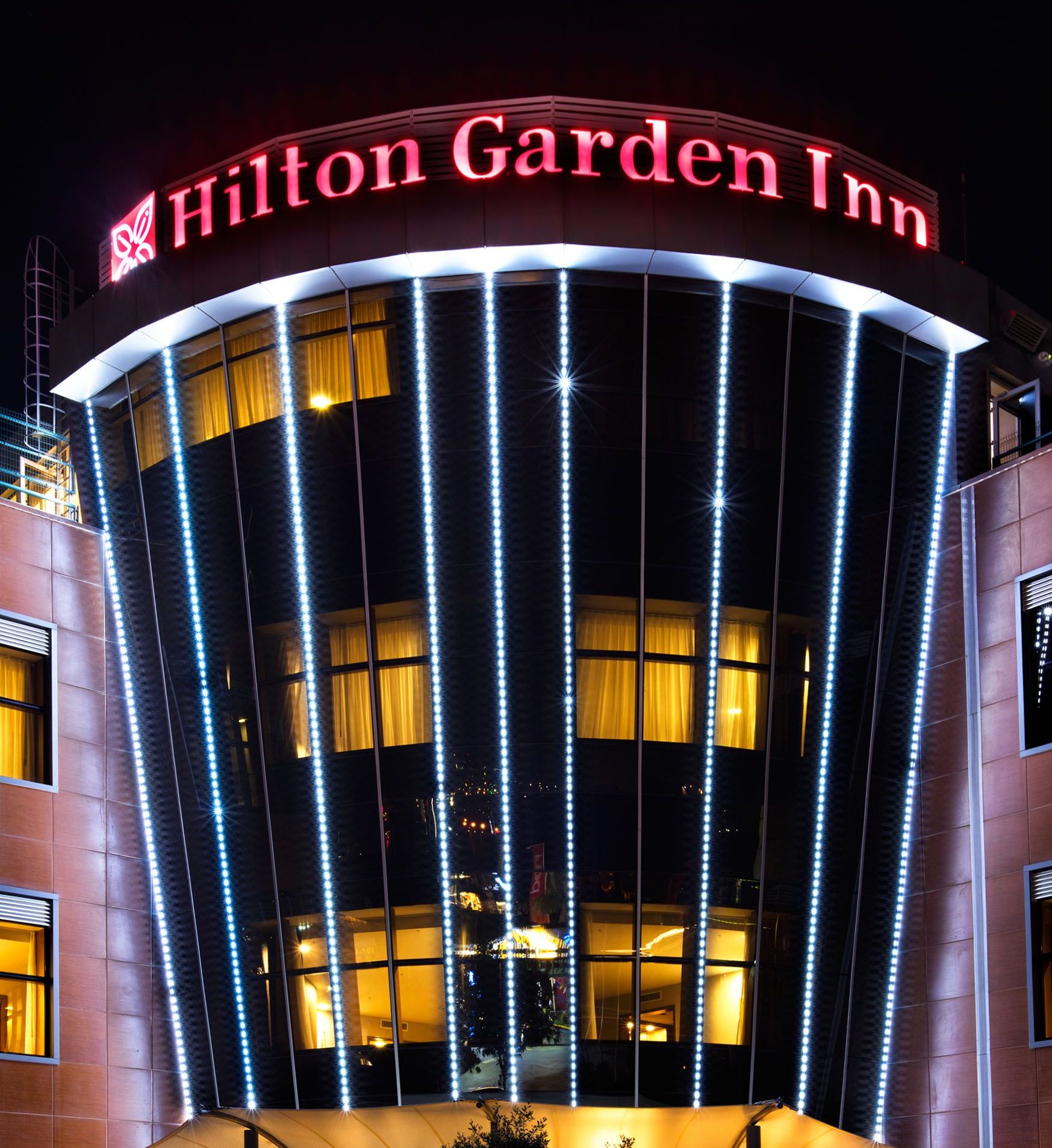 El exterior del Hilton Garden Inn con un letrero iluminado llamativo y franjas verticales de luz.