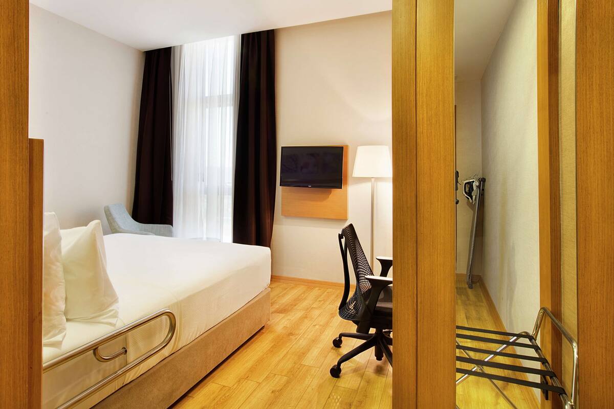 A hotel room with a bed, a desk, a chair, a TV, and a window with curtains.