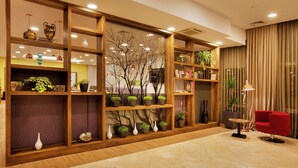 A lobby with wooden shelving displaying potted plants and decorative items, a red chair, and a table with a lamp.