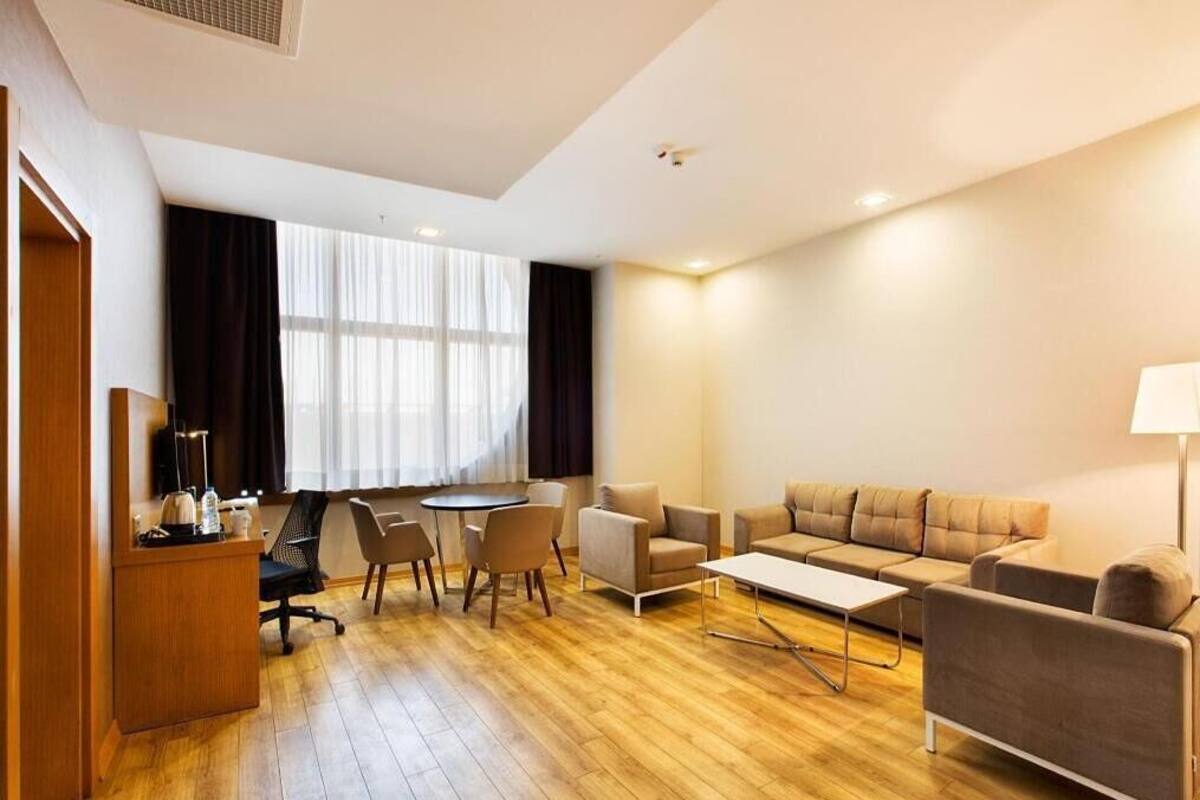 A modern hotel room with a wooden floor, a sofa set, a desk with a chair, and a large window with curtains.
