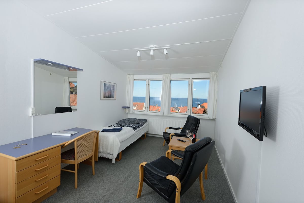 A compact bedroom with a single bed, a desk, a chair, a TV, and a window with a view of buildings and the sea.