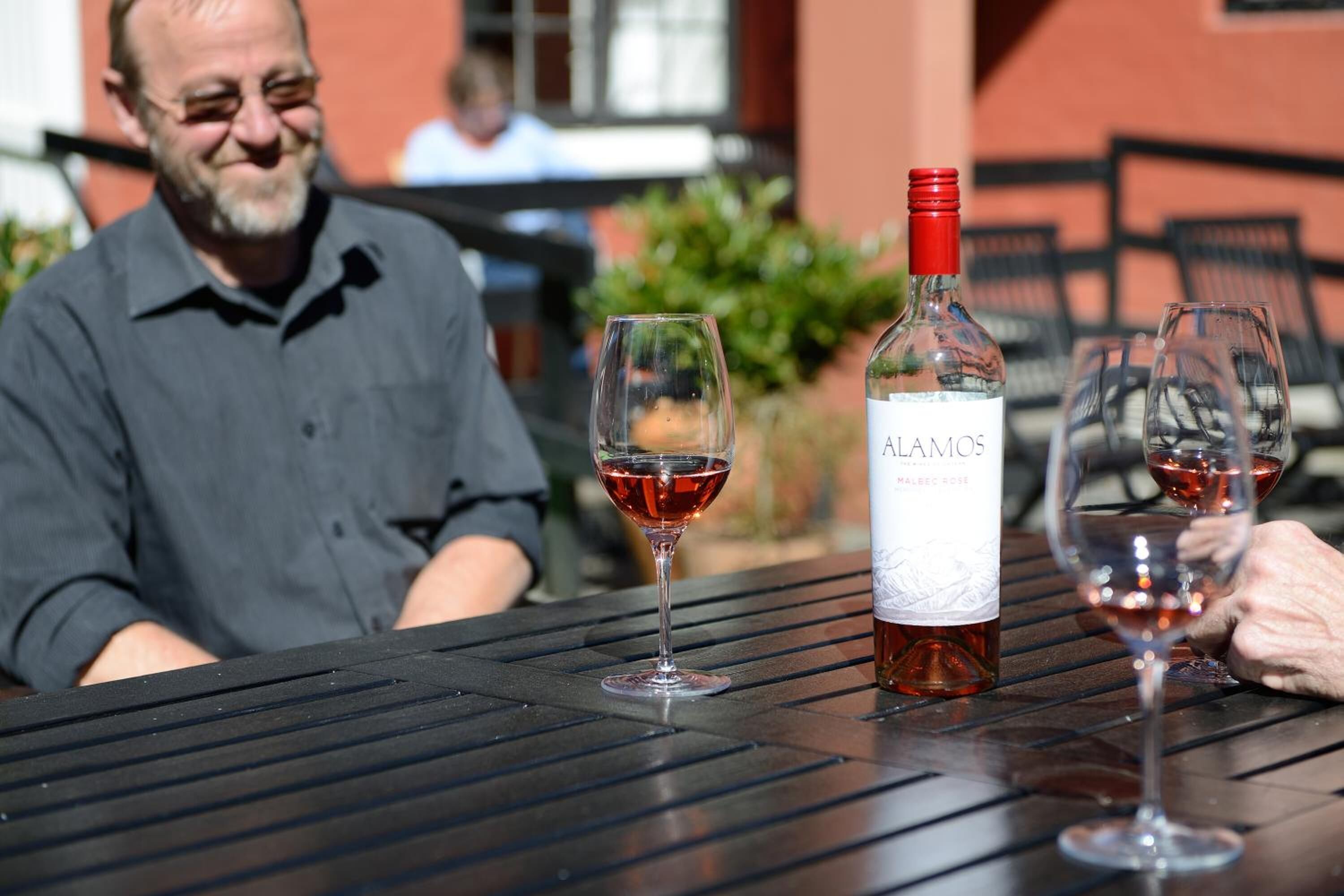 A table with a bottle of Alamos Malbec 2018 wine, two glasses of red wine, and a person seated in the background.