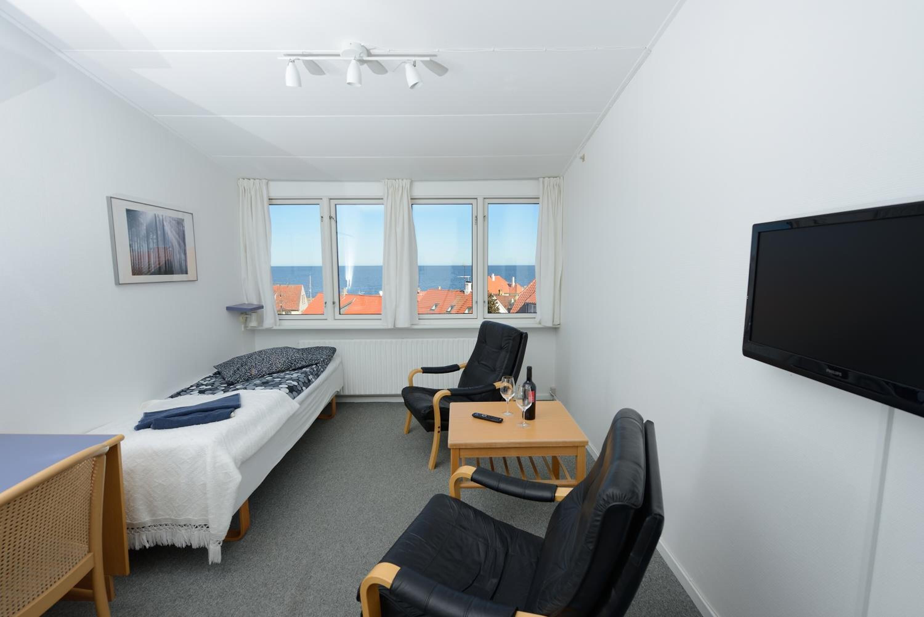 A room with a bed, a desk, a chair, a TV, and a window with a view of buildings and the sea.