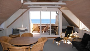 Un accogliente sottotetto con soffitto in legno, una TV, mobili in vimini e vista sul mare tramite una porta vetrata.