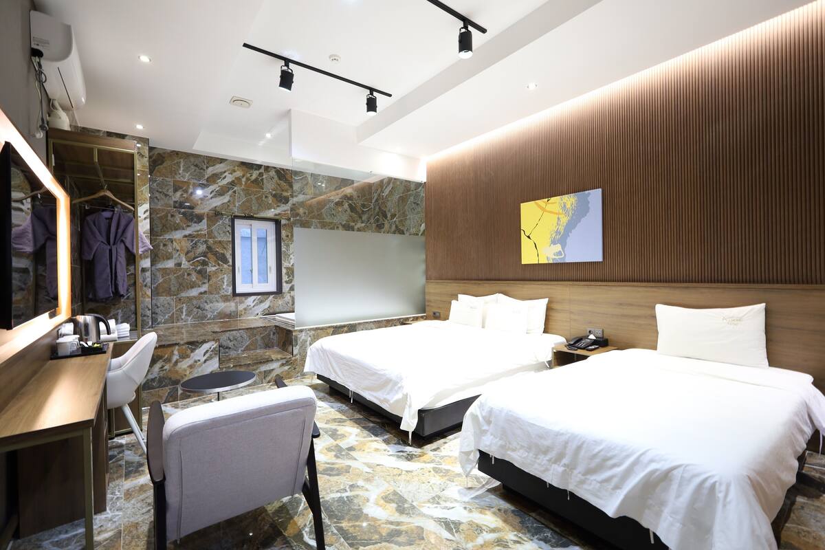A hotel room with two beds, a desk, a chair, a small round table, a robe rack, and a large mirror.